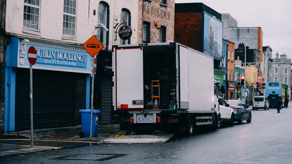 A white moving van is parked on a street in front of various shops, including one with a blue sign that reads 'Dublin Mouldings.' The van’s rear doors are open, revealing orange safety straps and eq