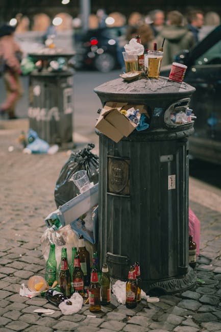 Indoor recycling area with a variety of cardboard boxes, paper packaging, and waste materials piled inside and on top of large blue and brown recycling bins. The backdrop features a blue sign with whi