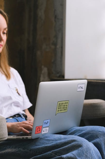 The image shows a young woman with long brown hair, sitting indoors on a grey sofa or armchair, working on a silver laptop placed on her lap. The laptop has several colorful stickers on its lid, inclu