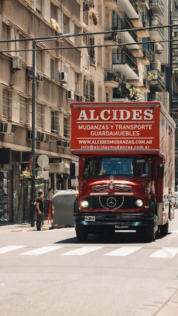 A red moving truck with the sign 'ALCIDES' on top, positioned on a city street in front of residential buildings with balconies and windows. The truck is part of a home relocation or furniture transpo