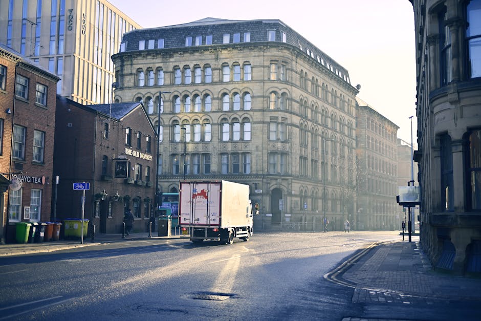 A red moving truck with the sign 'ALCIDES' on top, positioned on a city street in front of residential buildings with balconies and windows. The truck is part of a home relocation or furniture transpo